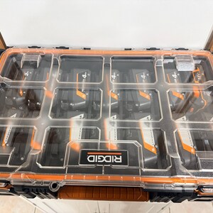 May include: A clear plastic and orange Ridgid tool organizer filled with black and orange 18V Lithium-Ion batteries. The organizer has multiple compartments and a handle for easy transport. The Ridgid logo is visible on the front.
