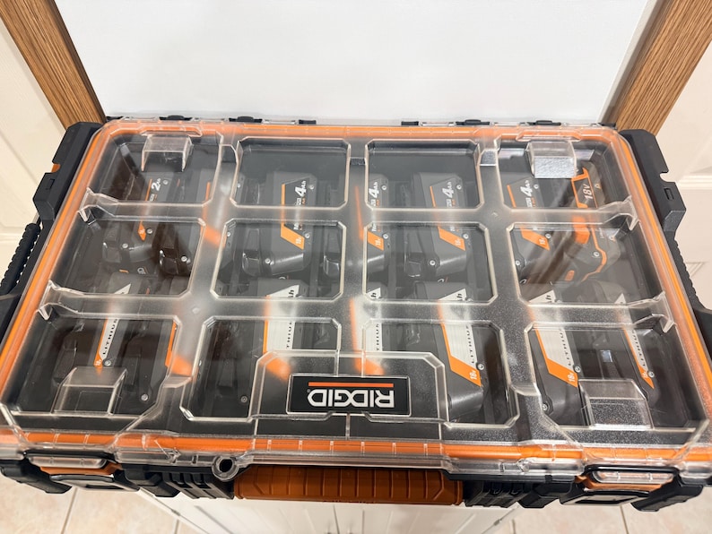 May include: A clear plastic and orange Ridgid tool organizer filled with black and orange 18V Lithium-Ion batteries. The organizer has multiple compartments and a handle for easy transport. The Ridgid logo is visible on the front.