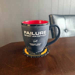 May include: Black coffee mug with the text "FAILURE IS NOT AN OPTION" and the NASA logo. The mug has a red interior and sits on a black and yellow coaster. The mug is from the Marshall Space Flight Center.