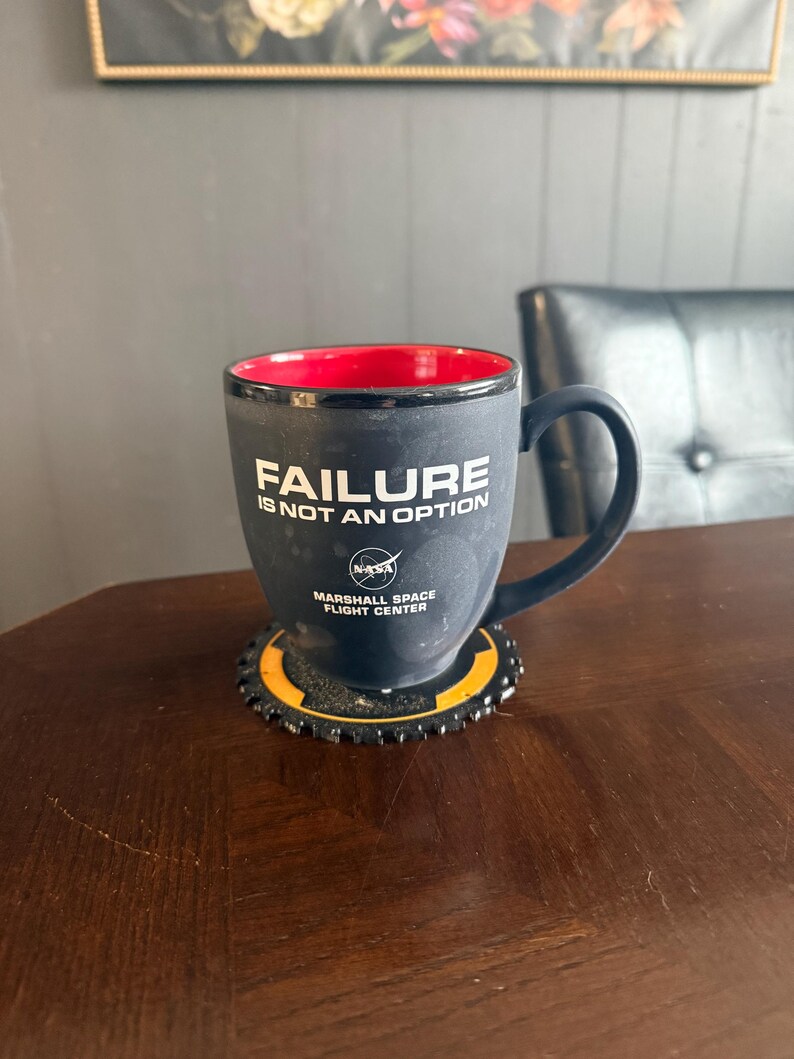 May include: Black coffee mug with the text "FAILURE IS NOT AN OPTION" and the NASA logo. The mug has a red interior and sits on a black and yellow coaster. The mug is from the Marshall Space Flight Center.