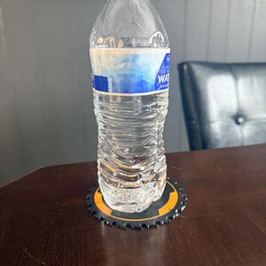 May include: A clear plastic water bottle with a white cap and a blue and white label, resting on a black and orange coaster with a gear-like design. The bottle is on a dark brown wooden table.