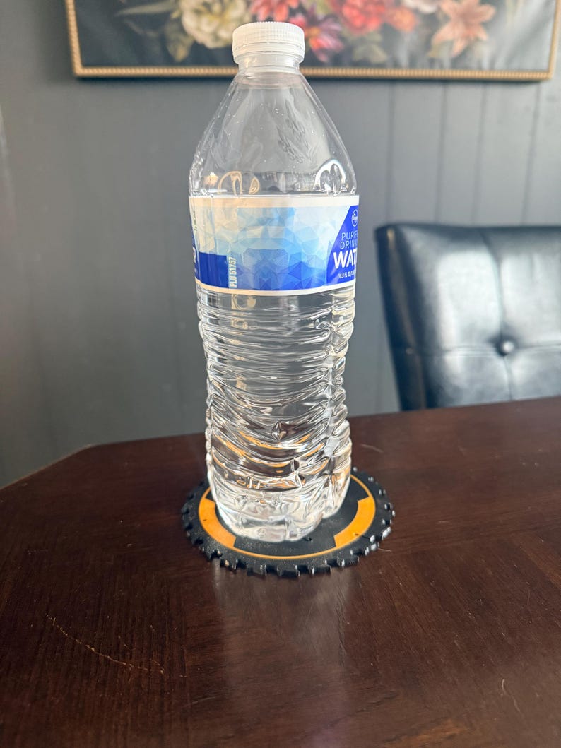 May include: A clear plastic water bottle with a white cap and a blue and white label, resting on a black and orange coaster with a gear-like design. The bottle is on a dark brown wooden table.