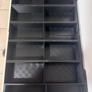May include: A black, multi-compartment organizer with a grid-like structure. The organizer features multiple rectangular compartments, arranged in rows and columns. The compartments are open-topped, providing easy access to stored items. The organizer is designed for storage and organization.