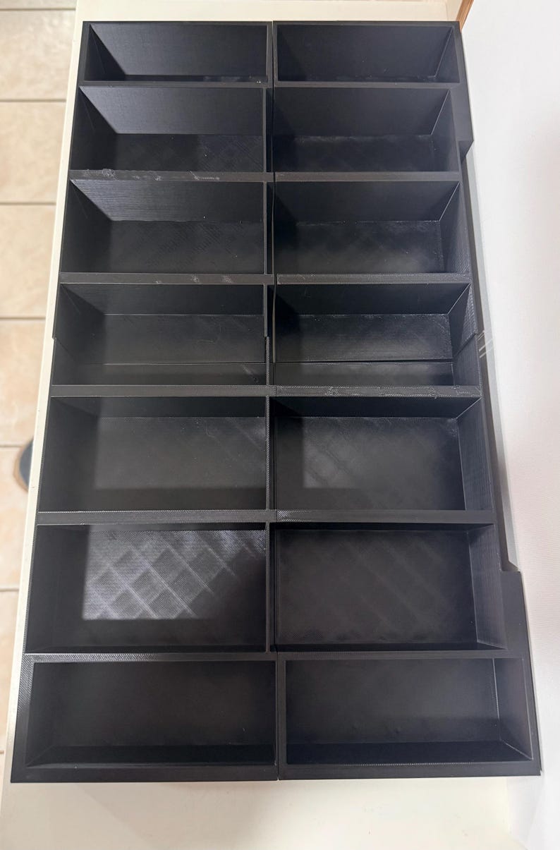 May include: A black, multi-compartment organizer with a grid-like structure. The organizer features multiple rectangular compartments, arranged in rows and columns. The compartments are open-topped, providing easy access to stored items. The organizer is designed for storage and organization.