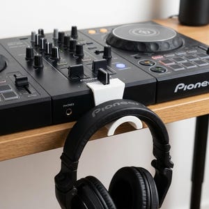 Peut inclure: Une table de mixage DJ noire avec un support de casque blanc et un casque noir. La table de mixage comporte de nombreux boutons, curseurs et boutons. Le casque est suspendu au support, qui est fixé à la table de mixage. Le mot "Pioneer" est visible.