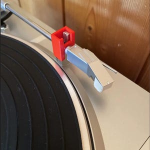 Może przedstawiać: Czerwony uchwyt ramienia 3D wydrukowany na srebrnym gramofonie. Ramię znajduje się nad czarną płytą winylową. Gramofon jest ustawiony na jasnobrązowym tle drewnianym.