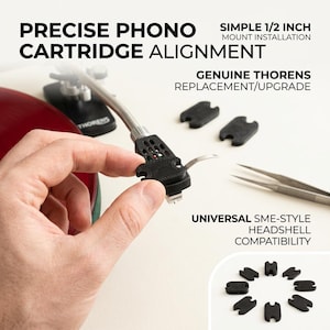 Peut inclure: Une cellule SME noire pour platine, tenue par une main. La cellule est étiquetée "CARRON" et est conçue pour un alignement précis de la cellule phono. L'image montre également des outils d'alignement noirs et le texte "GENUINE THORENS REPLACEMENT/UPGRADE."