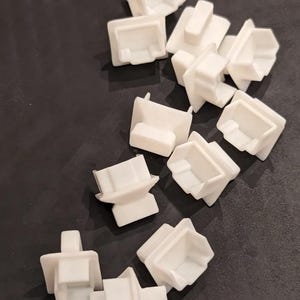 Può includere: Una collezione di piccoli angolari in plastica bianca, ciascuno con una base quadrata e una linguetta rettangolare sporgente. I pezzi sono sparsi su una superficie scura, mettendo in risalto il loro design geometrico e l'estetica minimalista.