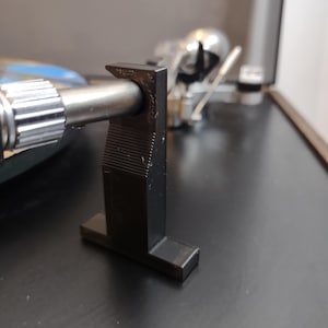 May include: Close-up of a black, L-shaped component of a turntable. The component is positioned on a dark surface, with a silver-coloured arm extending from it. The design features horizontal ridges on the vertical section.