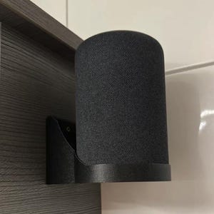 Op de afbeelding: Een zwarte, cilindervormige slimme luidspreker, gemonteerd op een zwarte wandbeugel. De luidspreker heeft een textuur die op stof lijkt en een afgeronde basis. De beugel is bevestigd aan een donkergrijze houten ondergrond.