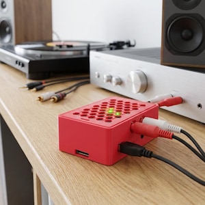 Könnte beinhalten: Ein rechteckiges rotes elektronisches Gerät mit einer wabenförmigen Oberseite und zwei leuchtend grünen Kontrollleuchten. Das Gerät ist über rote, weiße und schwarze Kabel mit einem silbernen Audioverstärker verbunden und steht auf einer Holzoberfläche.