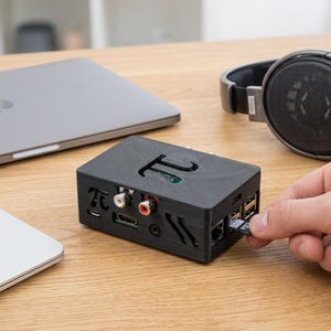 Puede incluir: Una carcasa de ordenador rectangular negra con un recorte del símbolo pi en la parte superior. La carcasa cuenta con puertos laterales que incluyen dos conectores RCA, un puerto HDMI y puertos USB. Se inserta una tarjeta de memoria pequeña.