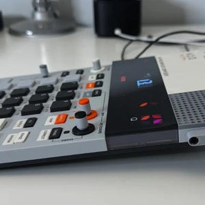 Op de afbeelding: Een grijs elektronisch muziekproductieapparaat met zwarte en oranje knoppen, knoppen en een beeldscherm. Het apparaat heeft een raster van zwarte knoppen en een ingebouwde standaard. Er is een witte kaart bovenop geplaatst.