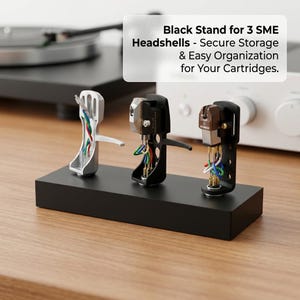Peut inclure: Un support noir présentant trois cellules SME. Les cellules sont argentées, noires et marron, chacune avec des fils colorés. Le support est rectangulaire et repose sur une surface en bois. Le texte sur l'image indique : Black Stand for 3 SME Headshells - Secure Storage & Easy Organization for Your Cartridges.