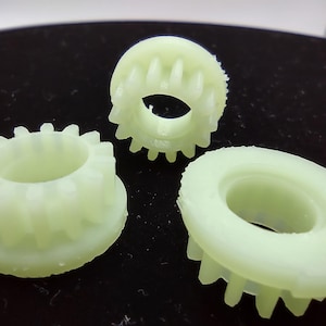 May include: Three pale green plastic gears with toothed edges. The gears have a central hole and varying widths. The gears are likely components for a mechanical device or model.