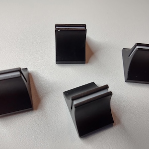 Op de afbeelding: Vier zwarte kastknoppen met een gebogen ontwerp en een dunne, lichtgekleurde accentlijn. De knoppen zijn vierkant en hebben een moderne, minimalistische uitstraling, geschikt voor laden of deuren.