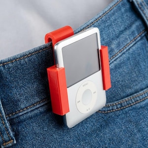 May include: A silver iPod music player secured in a bright red holder clipped to the waistband of blue jeans. The iPod has a screen and a circular control pad with a menu button. The holder is made of plastic.