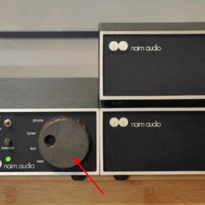 May include: Black audio components with silver trim, featuring large, textured knobs. The units are stacked, with the brand name "naim audio" visible. A green light indicates power. The overall design is minimalist and modern.