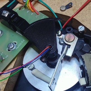 May include: Detailed view of electronic components, featuring circuit boards, wires, and metal elements. A black plastic part and a silver metal piece are visible. Various coloured wires connect to the components, set against a light brown background.