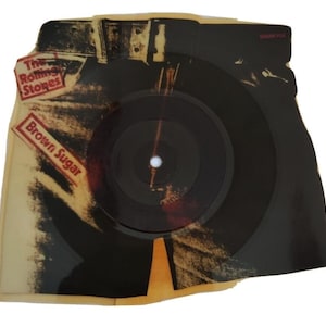 Può includere: Un disco in vinile vintage con un disco nero e una custodia a forma di jeans. La custodia presenta il testo "The Rolling Stones" e "Brown Sugar" in rosso e bianco. Il disco è un oggetto da collezione.