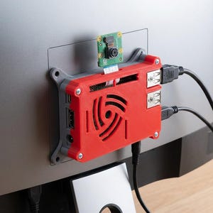 Pode incluir: Uma caixa de plástico vermelha e cinzenta que aloja um computador de placa única, montada na parte de trás de um monitor. A caixa apresenta uma ventoinha circular, um pequeno módulo de câmara com cabo plano e vários cabos pretos ligados.