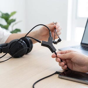 May include: A black audio splitter with three ports is connected to headphones, earbuds, and a smartphone. The splitter is held by a person. A pair of black over-ear headphones are on a light wood surface.