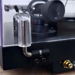 Può includere: Primo piano di un giradischi nero con una copertura in plastica trasparente. Il giradischi presenta un braccio argentato e una leva di sollevamento in plastica trasparente. I jack audio dorati e rossi sono visibili sul retro.