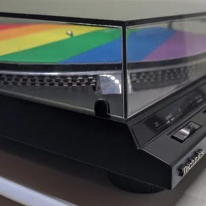May include: A black Technics turntable with a rainbow-colored design on the lid. The turntable features a silver platter and control buttons. The design includes the colors red, orange, yellow, green, blue, and purple.