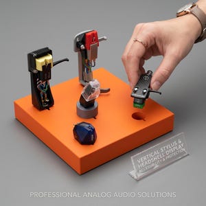 Peut inclure: Un présentoir orange avec plusieurs cellules et pointes de lecture de platine vinyle noires et argentées. Une main tient une des cellules. Le présentoir porte une pancarte en acrylique transparent avec l'inscription "VERTICAL STYLUS & HEADSHELL DISPLAY ACCESSIBILITY & PROTECTION."