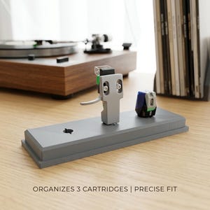 Könnte beinhalten: Ein grauer Patronenhalter mit zwei Tonabnehmern. Der Halter trägt den Text "ORGANIZES 3 CARTRIDGES | PRECISE FIT". Im Hintergrund befindet sich ein Plattenspieler aus Holz.