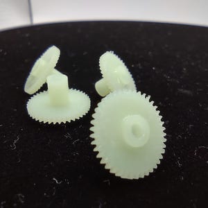 May include: Several small, off-white plastic gears with toothed edges are scattered on a dark surface. The gears vary in size and have a central hole. The lighting highlights the gear teeth and the overall shape of the components.