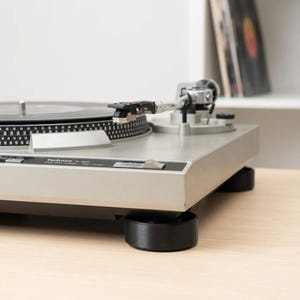 Könnte beinhalten: Silberner Technics SL-1200 Plattenspieler mit schwarzer Schallplatte. Der Tonarm befindet sich über der Platte. Der Plattenspieler steht auf schwarzen zylindrischen Stützen. Vinylplatten sind auf einem Regal im Hintergrund zu sehen.