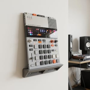 Op de afbeelding: Een grijze en witte K.O. II 64 MB Sampler Composer aan de muur. Het apparaat heeft een scherm, knoppen en een luidspreker. De tekst "K.O. II" en Japanse karakters zijn zichtbaar. Het is gemonteerd op een grijze plank.