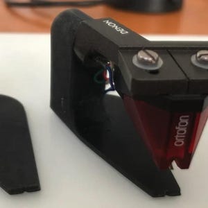 Könnte beinhalten: Schwarzer Denon-Plattenspieler-Tonabnehmer mit roter Ortofon-Nadel. Der Tonabnehmer ist auf einem schwarzen Headshell montiert. Links befindet sich ein schwarzer Ständer. Audiogeräte.