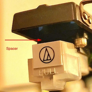 Peut inclure: Gros plan sur une entretoise de cartouche Audio-Technica blanche et grise. L'entretoise porte un logo noir et le mot "PULL" imprimé. La cartouche est fixée à un composant noir.