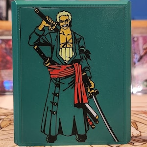 May include: Green card storage box featuring a character illustration with swords and a red sash. The box has a dark green border and a detailed graphic design. The background includes other card storage boxes.