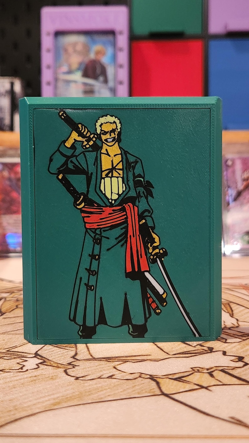 May include: Green card storage box featuring a character illustration with swords and a red sash. The box has a dark green border and a detailed graphic design. The background includes other card storage boxes.