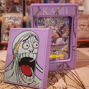 May include: A lilac card box with gold lettering spelling "KAMI". Inside is a trading card with a vibrant illustration. A matching card is on the left, showing a character's face with an open mouth and a shocked expression.