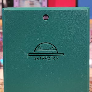 May include: A teal-colored card case with a textured surface. The case features a hat design and the text "SKERPOTCG" in white. The background includes other colorful card cases.