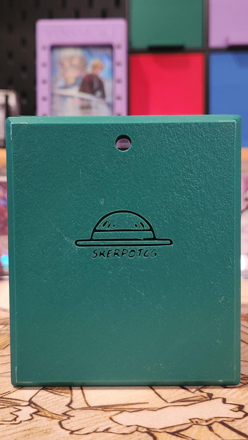 May include: A teal-colored card case with a textured surface. The case features a hat design and the text "SKERPOTCG" in white. The background includes other colorful card cases.