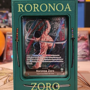 May include: A green card holder featuring a card with the name "RORONOA" at the top. The card inside depicts a muscular character with swords. The text "ZORO" is at the bottom.