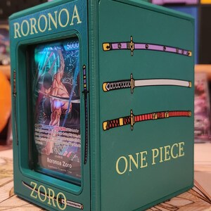 May include: Green box featuring a Roronoa Zoro trading card from the One Piece series. The box has the name "RORONOA ZORO" and "ZORO" printed on it. The box is decorated with three katana sword illustrations and the words "ONE PIECE".