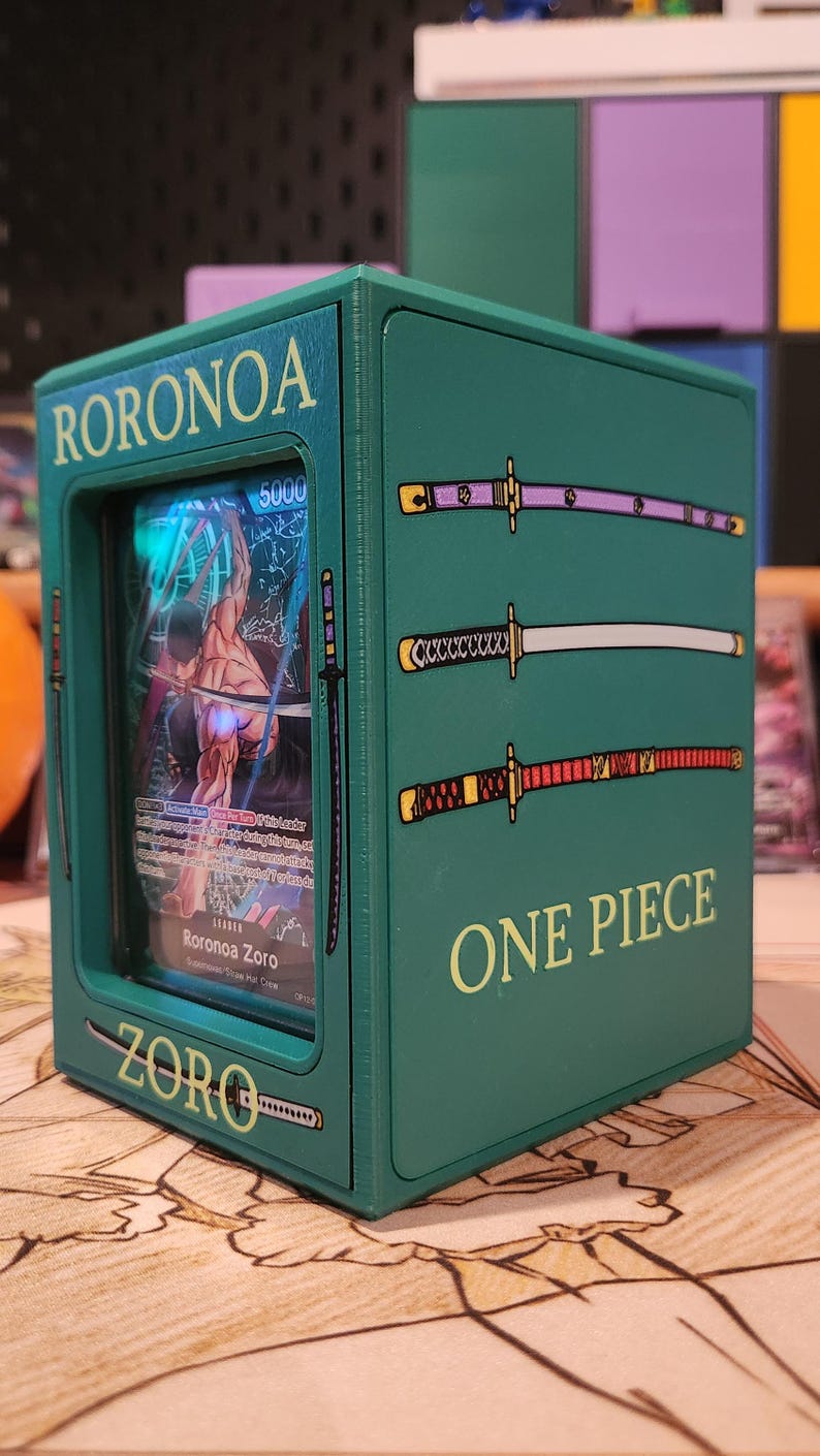 May include: Green box featuring a Roronoa Zoro trading card from the One Piece series. The box has the name "RORONOA ZORO" and "ZORO" printed on it. The box is decorated with three katana sword illustrations and the words "ONE PIECE".
