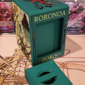 May include: A teal card storage box with the name "RORONOA ZORO" printed on it. The box has a matching lid with a circular handle. The box is decorated with illustrations of a character with swords.