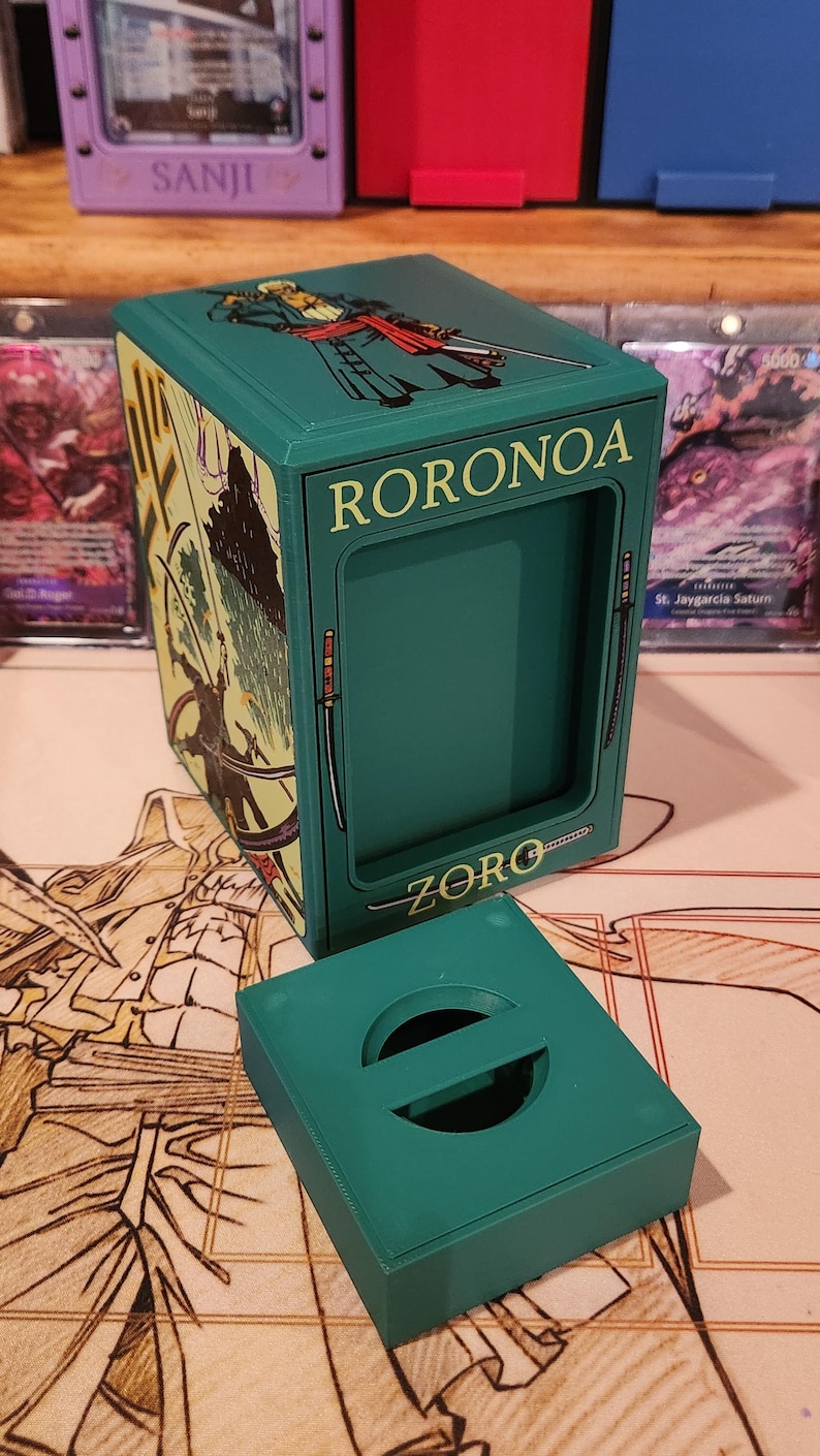 May include: A teal card storage box with the name "RORONOA ZORO" printed on it. The box has a matching lid with a circular handle. The box is decorated with illustrations of a character with swords.