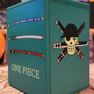 May include: A teal box featuring the "One Piece" logo and imagery. The box has three swords on one side and a skull and cross swords on another. The text "ONE PIECE" is printed on the front of the box.