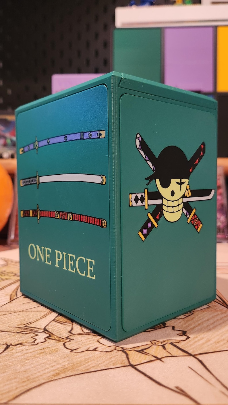 May include: A teal box featuring the "One Piece" logo and imagery. The box has three swords on one side and a skull and cross swords on another. The text "ONE PIECE" is printed on the front of the box.