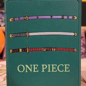 May include: A teal card holder featuring three stylized katana swords in purple, silver, and red. The text "ONE PIECE" is printed in yellow at the bottom of the holder. The design is a reference to the popular anime series.
