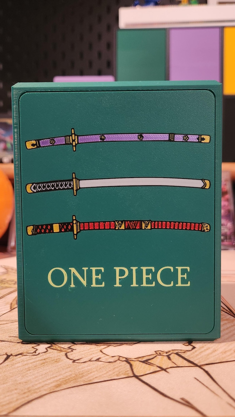 May include: A teal card holder featuring three stylized katana swords in purple, silver, and red. The text "ONE PIECE" is printed in yellow at the bottom of the holder. The design is a reference to the popular anime series.