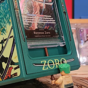 May include: A green card holder with the name "RORONOA" at the top and "ZORO" at the bottom. Inside the holder is a card featuring a character named Roronoa Zoro. A small Lego figure of Zoro is holding a sword. The card holder has a sword design.